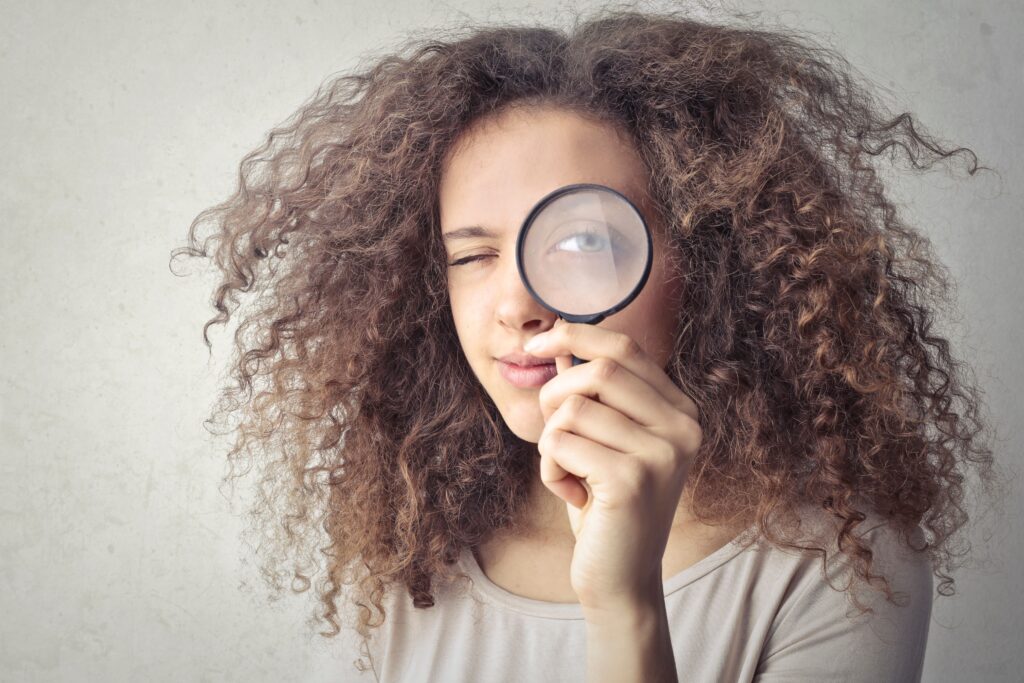 Frau mit braunem lockigem Haar guckt durch eine Lupe
