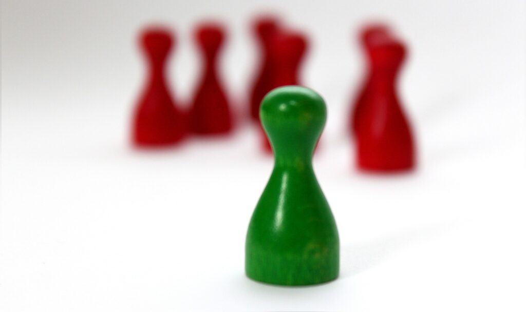 Eine grüne Spielfigur aus Holz steht mit sechs roten Spielfiguren im Hintergrund stehend
